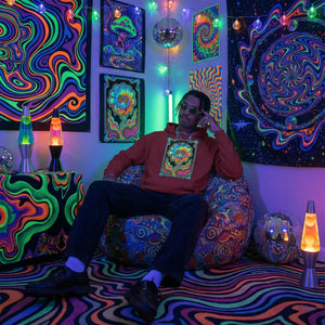 Man wearing red psychedelic hoodie sitting on patterned bean bag in neon art room with lava lamps and disco balls