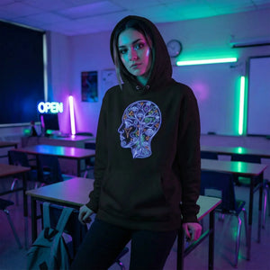 Young woman in black hoodie with neon psychedelic face design, standing in neon-lit classroom with 'OPEN' sign