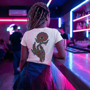 Person with dreadlocks wearing a white psychedelic t-shirt with an eye-flower print, leaning on neon-lit bar