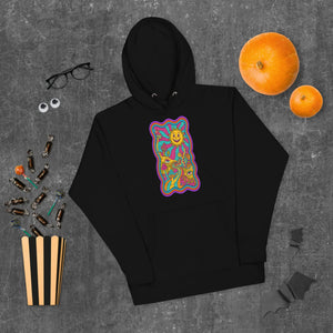 Black unisex premium pullover hoodie with colorful psychedelic sun and mushrooms design, flat lay with Halloween candy, pumpkins, glasses, and googly eyes