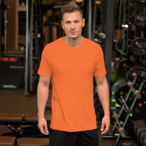 Man wearing plain orange unisex staple t-shirt standing in gym with workout equipment