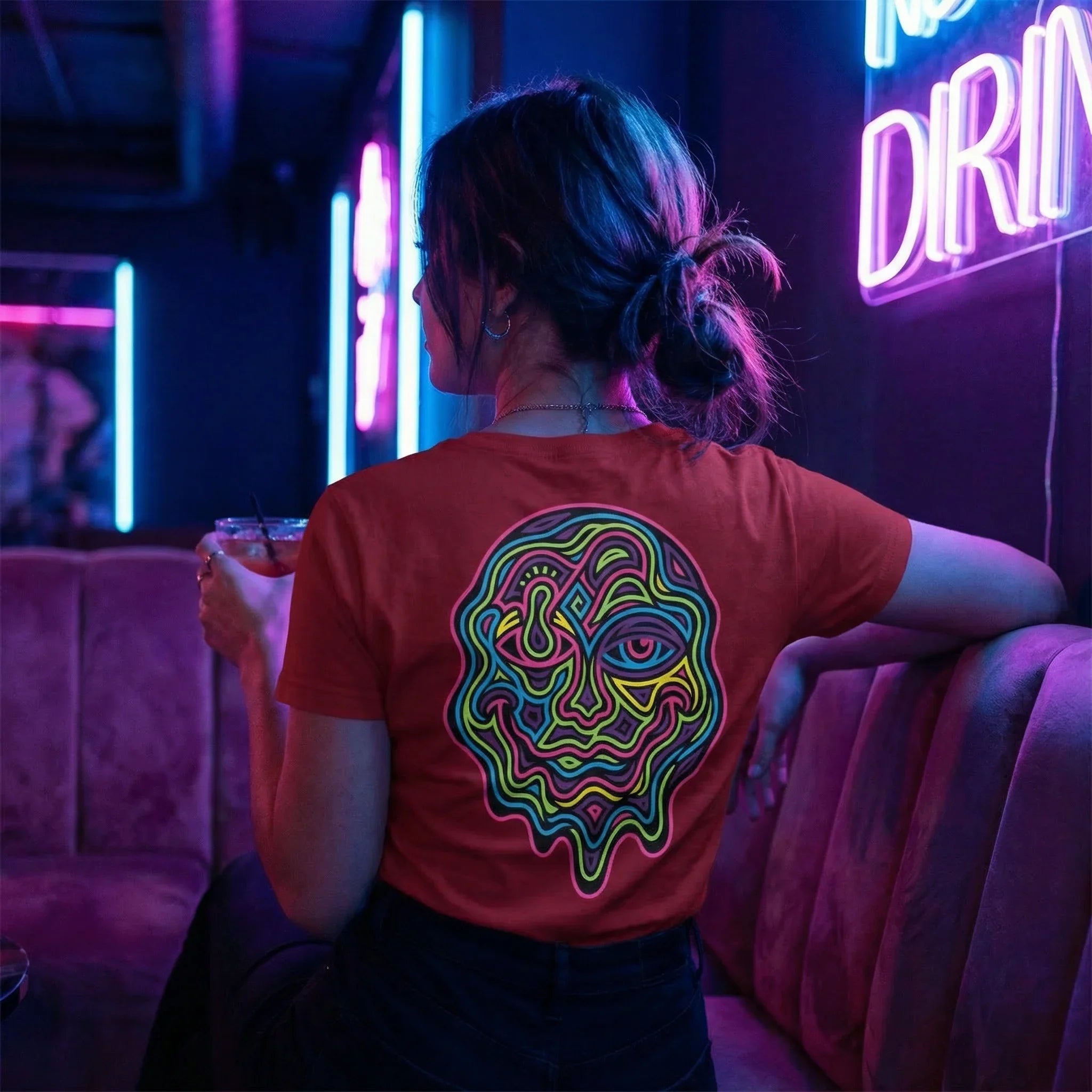 Woman wearing red t-shirt with colorful psychedelic face design back, sitting in neon-lit bar holding drink