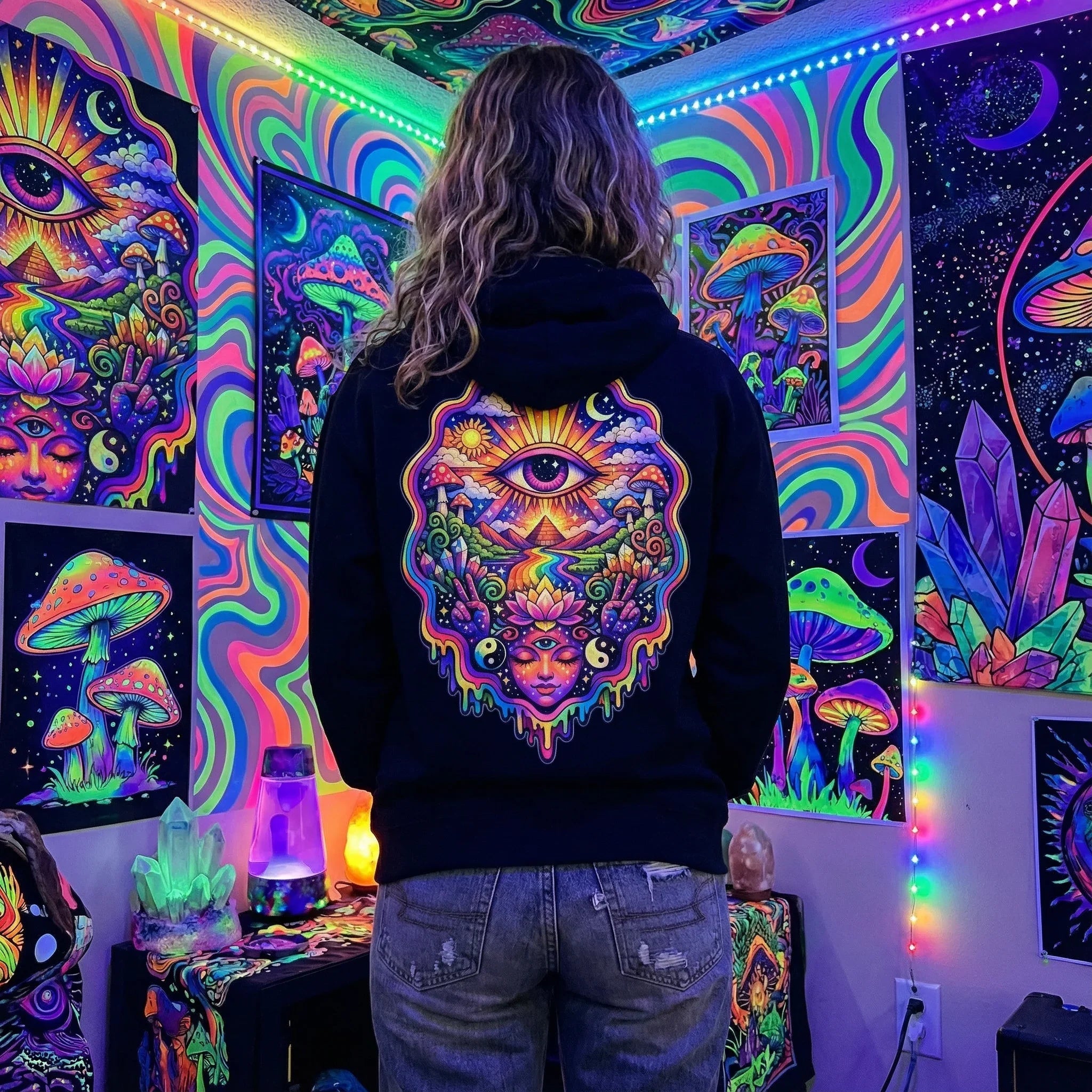 Person wearing black hoodie with colorful psychedelic eye and mushroom design, surrounded by neon art posters and crystals