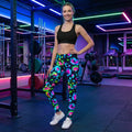 Smiling woman in bright psychedelic leggings and black sports bra posing in neon-lit gym with workout equipment