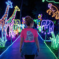 Woman wearing pink tiger t-shirt walking through neon animal light display with giraffes, elephants, and monkey