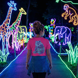 Woman wearing pink tiger t-shirt walking through neon animal light display with giraffes, elephants, and monkey