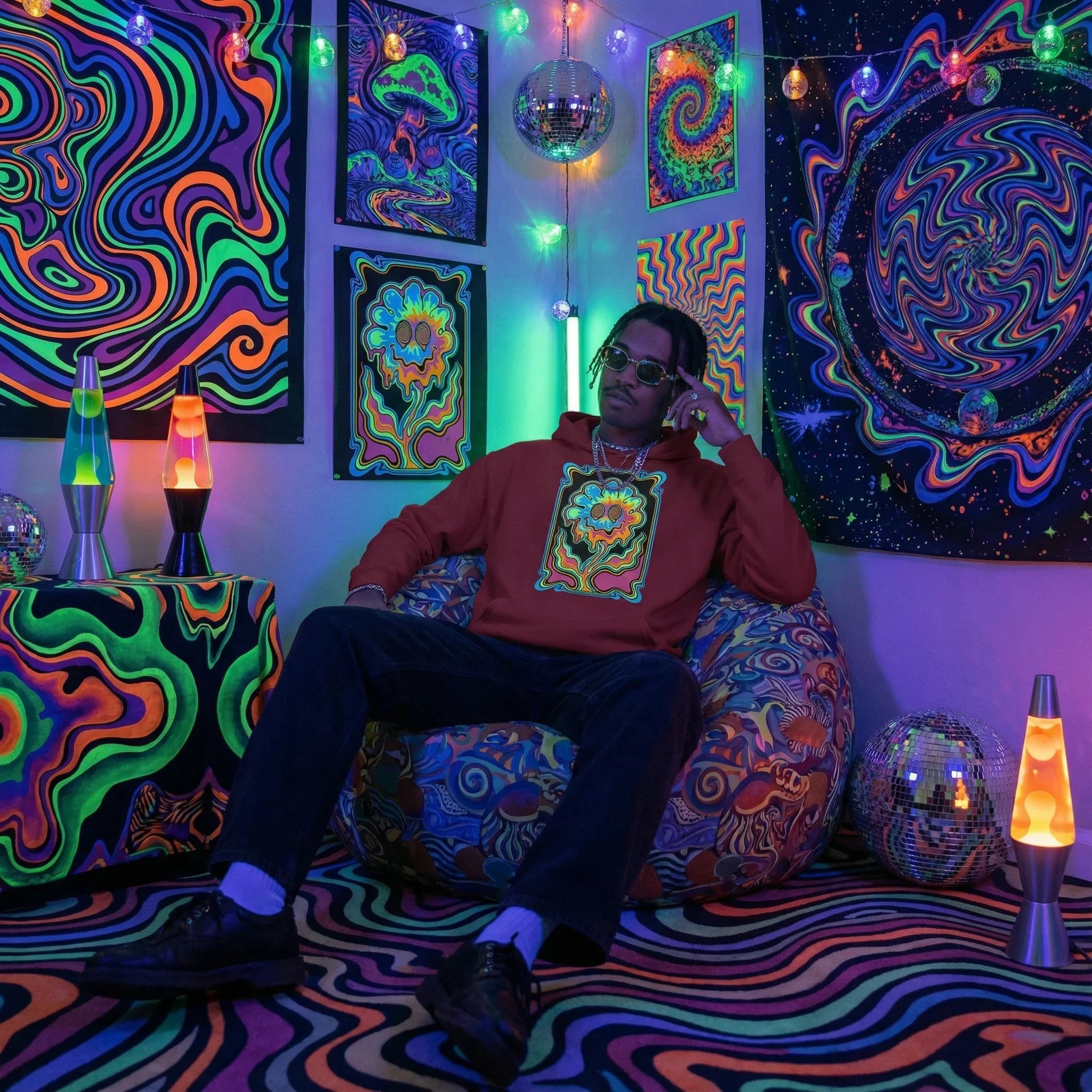 Man wearing red psychedelic hoodie sitting on patterned bean bag in neon art room with lava lamps and disco balls