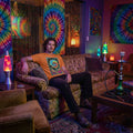 Man in psychedelic orange T-shirt with colorful skull design sitting on vintage sofa in neon-lit room with tie-dye tapestries and lava lamps
