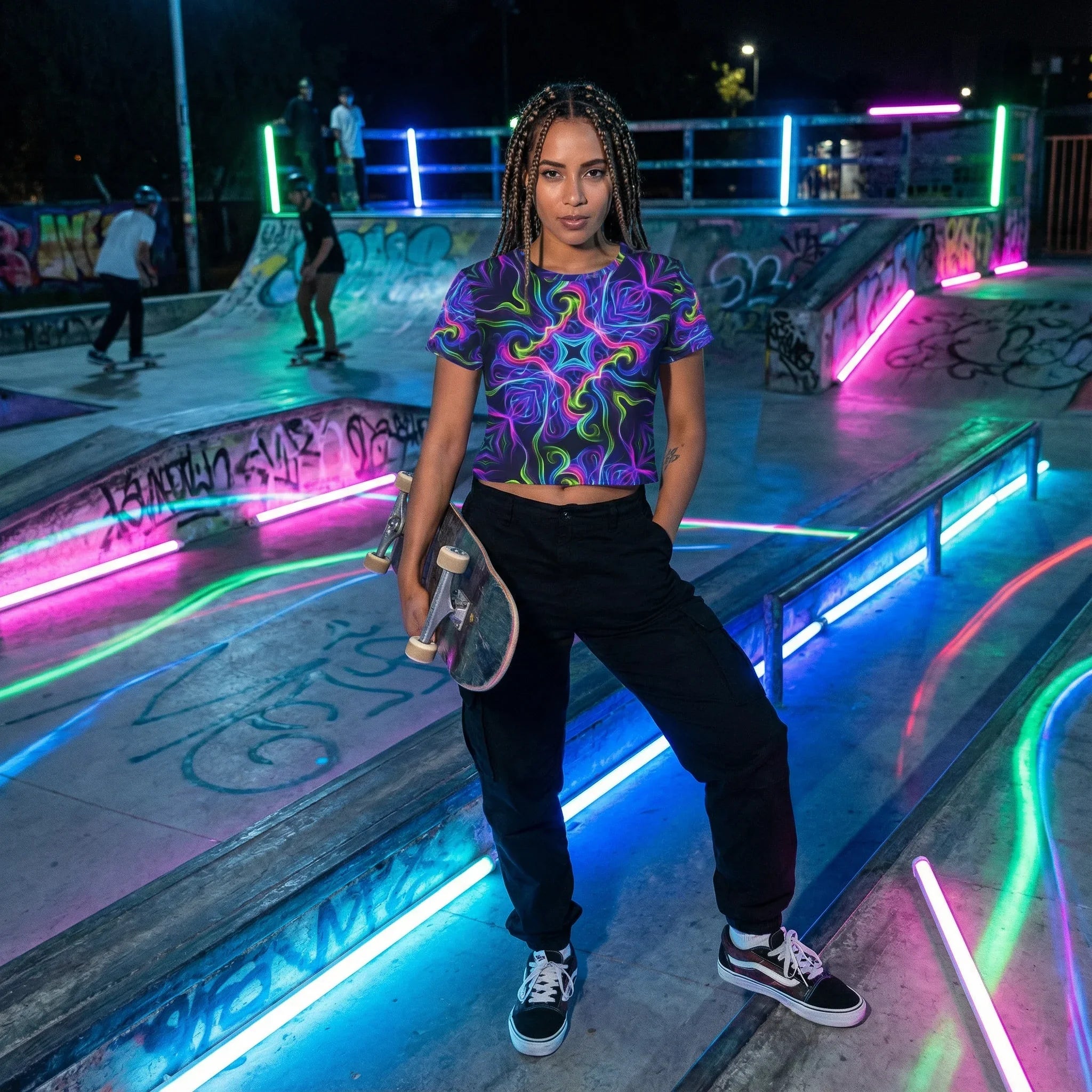 Young woman with braided hair wearing a vibrant psychedelic crop top and black pants holding a skateboard at a neon-lit graffiti skatepark at night