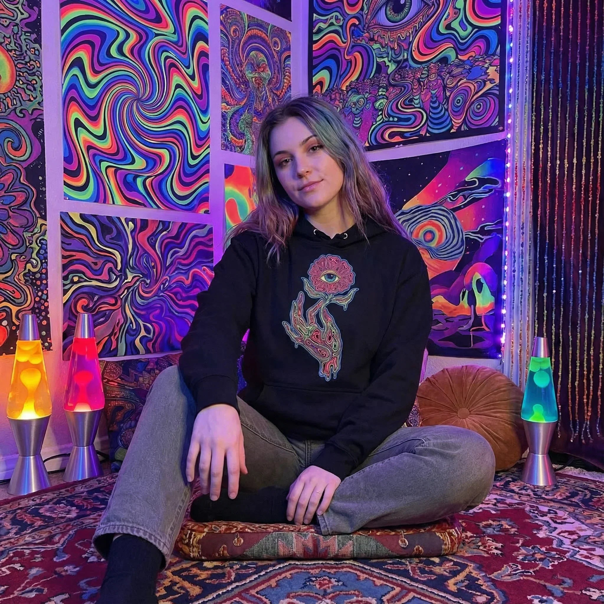 Woman sitting cross-legged on a patterned rug in a psychedelic art room wearing black hoodie with eye and hand design, surrounded by colorful lava lamps
