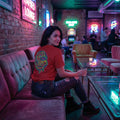 Woman wearing red psychedelic graphic T-shirt and ripped jeans sitting on velvet sofa in neon-lit lounge bar