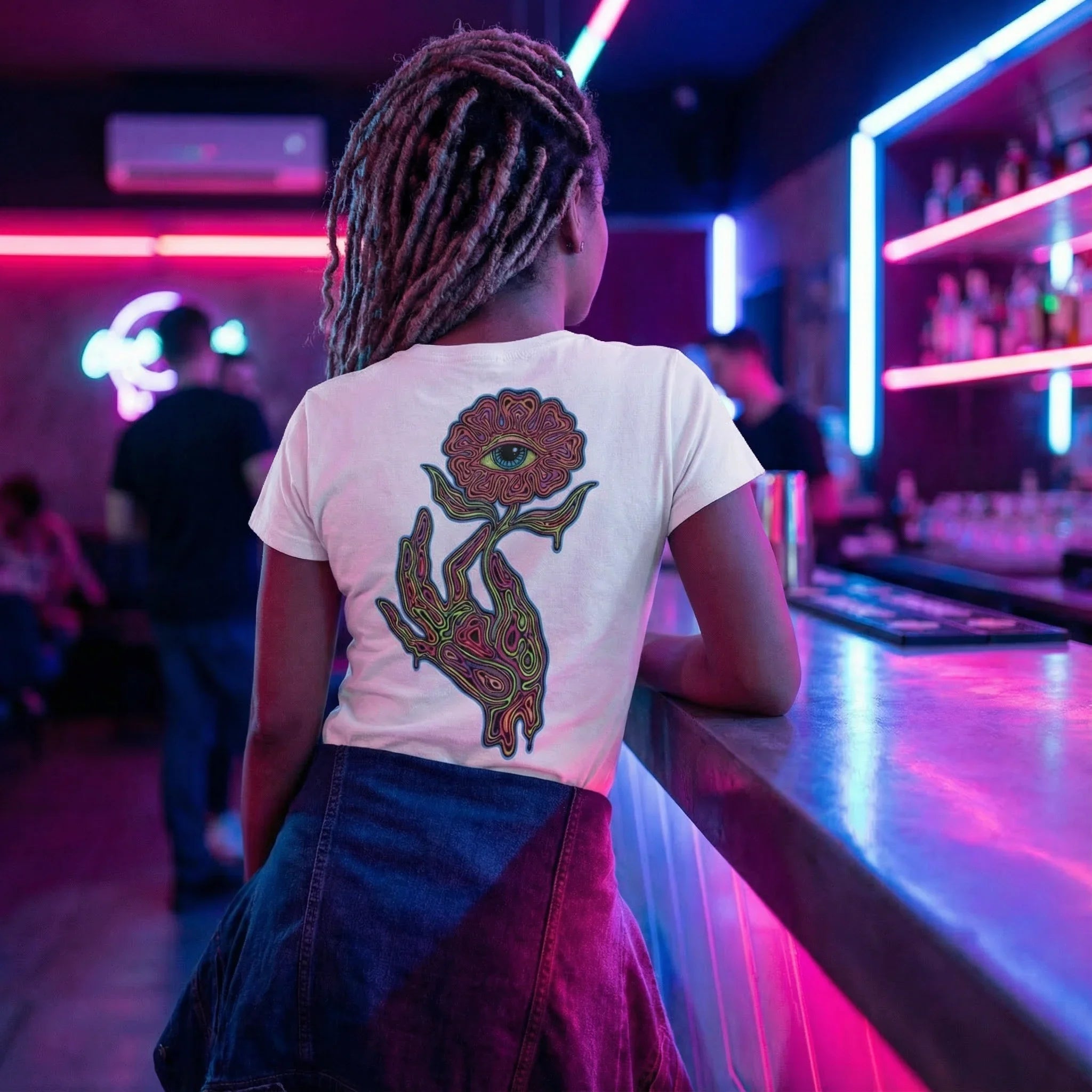 Person with dreadlocks wearing a white psychedelic t-shirt with an eye-flower print, leaning on neon-lit bar