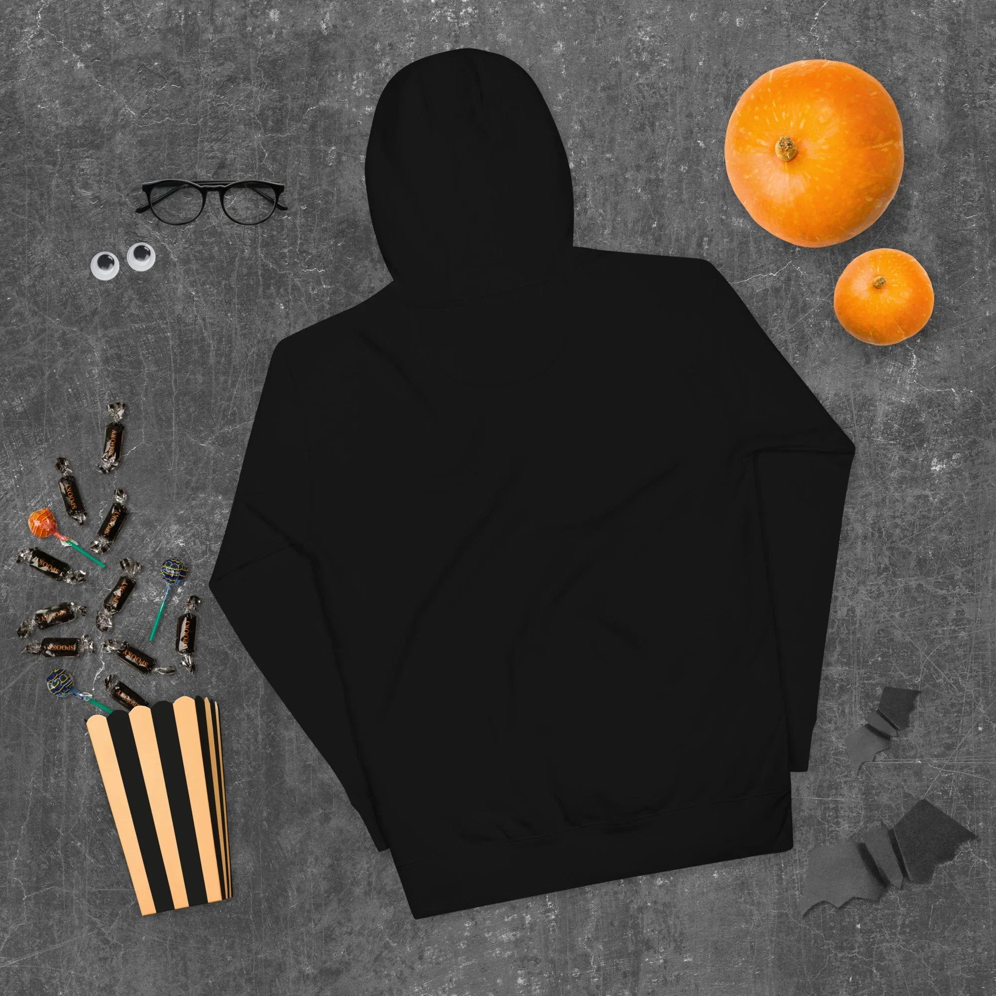 Black unisex premium pullover hoodie on dark textured surface with orange pumpkins, glasses, and Halloween candy