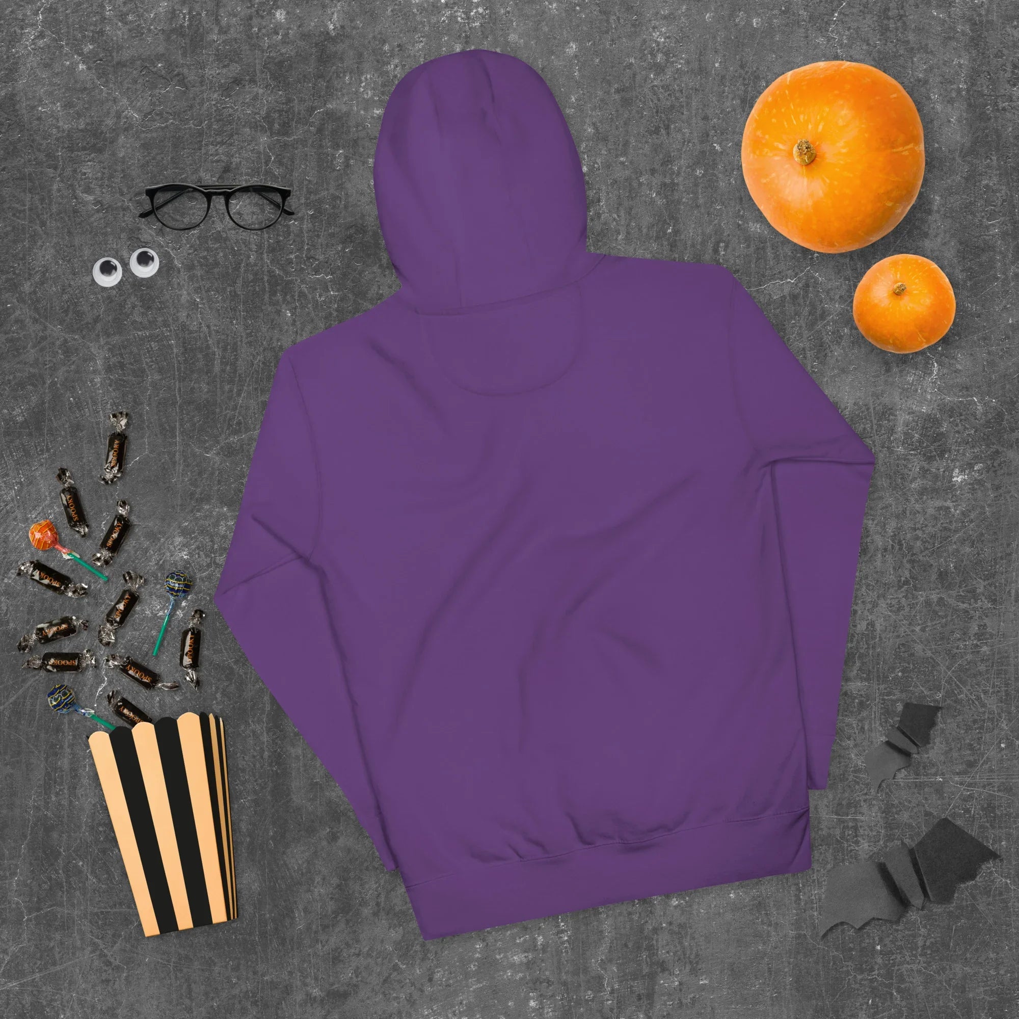 Purple unisex premium pullover hoodie laid flat on gray surface with Halloween pumpkins, candy, glasses, and paper bats