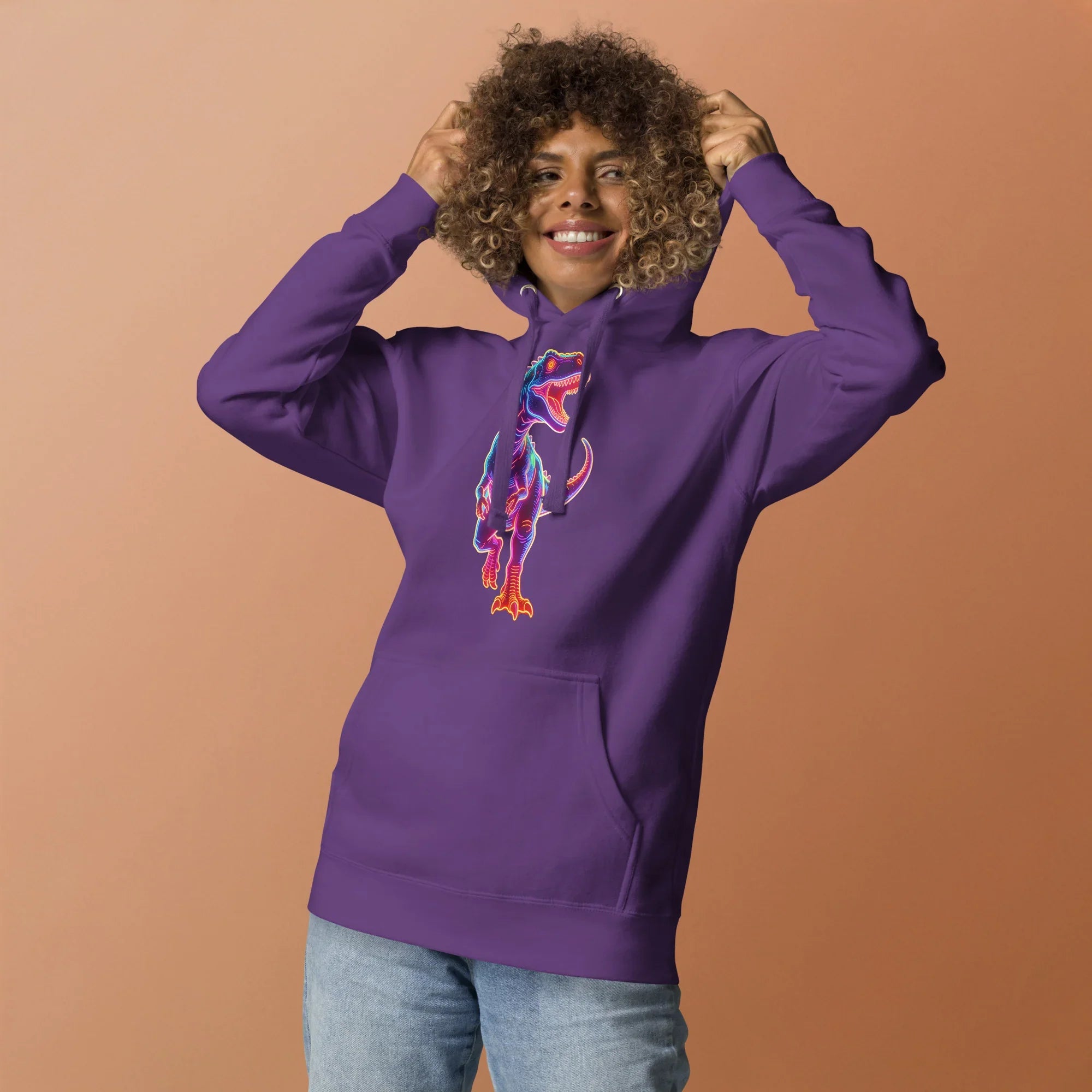 Smiling woman wearing purple hoodie with neon dinosaur graphic, light blue jeans, beige background