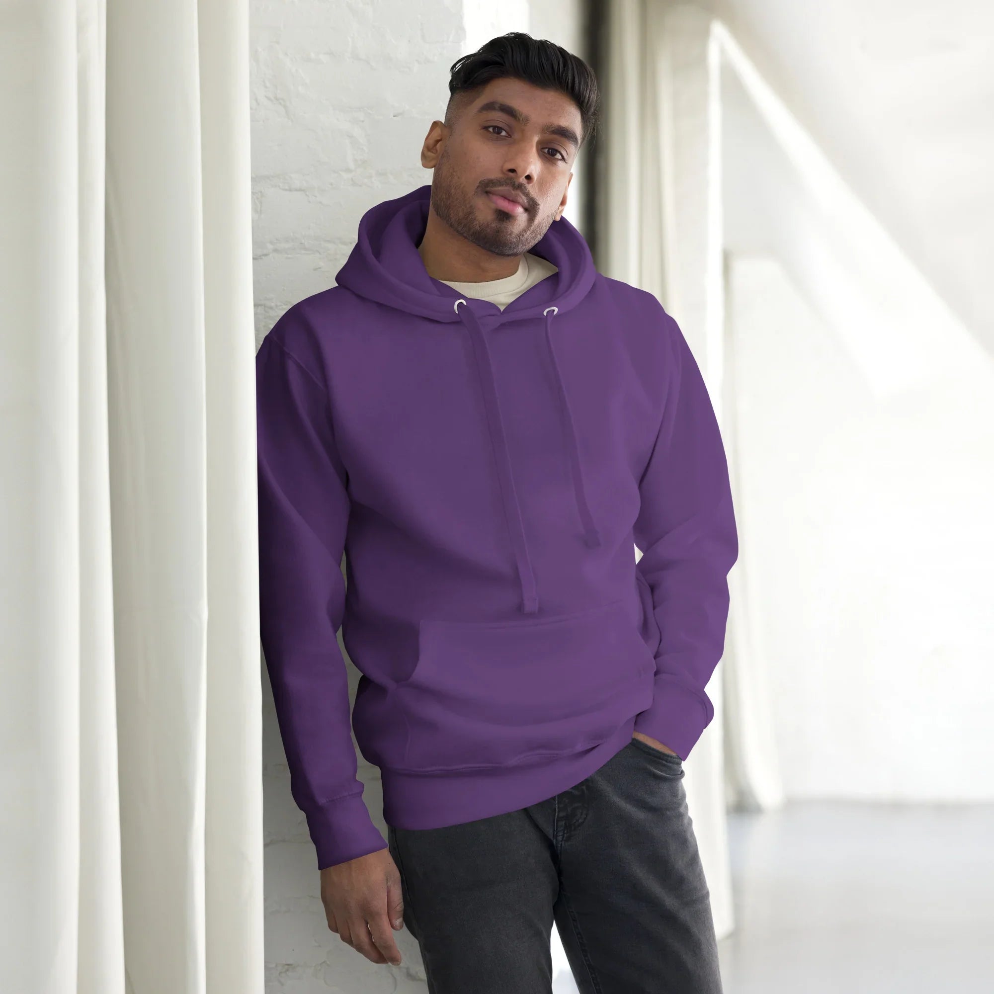 Man wearing a purple unisex premium pullover hoodie with front pocket, standing indoors by white curtains
