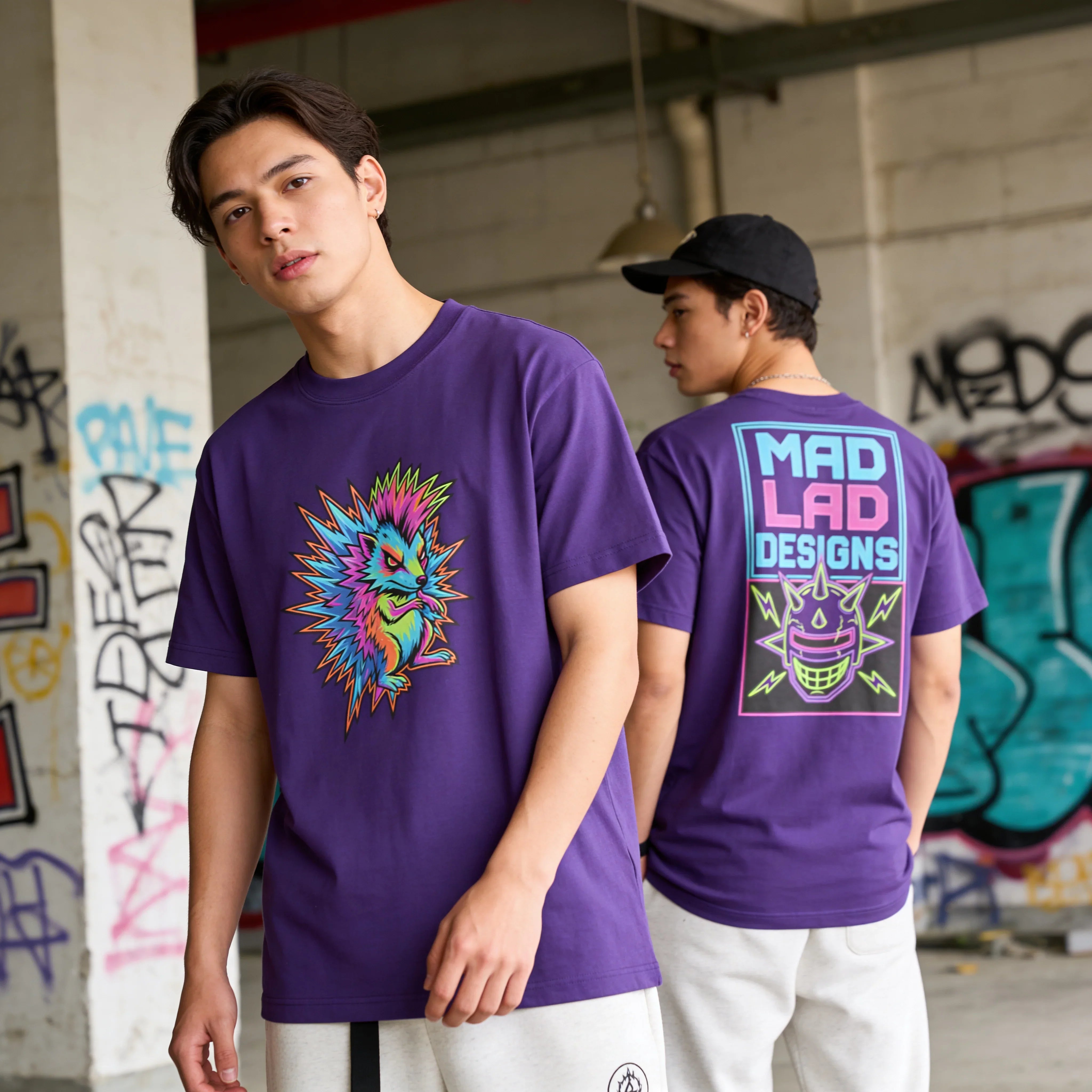 Men modeling Mad Lad Designs purple t-shirts with neon psychedelic hedgehog and logo in urban graffiti setting.