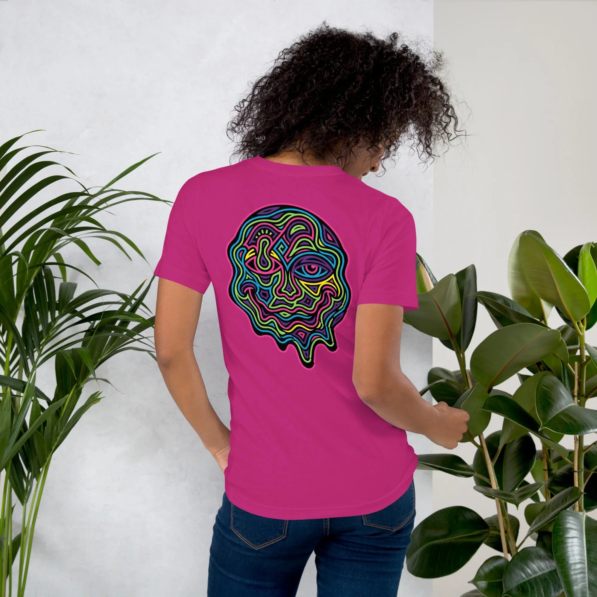 Woman wearing berry-colored unisex t-shirt with vibrant psychedelic neon abstract face design on back, green plants background