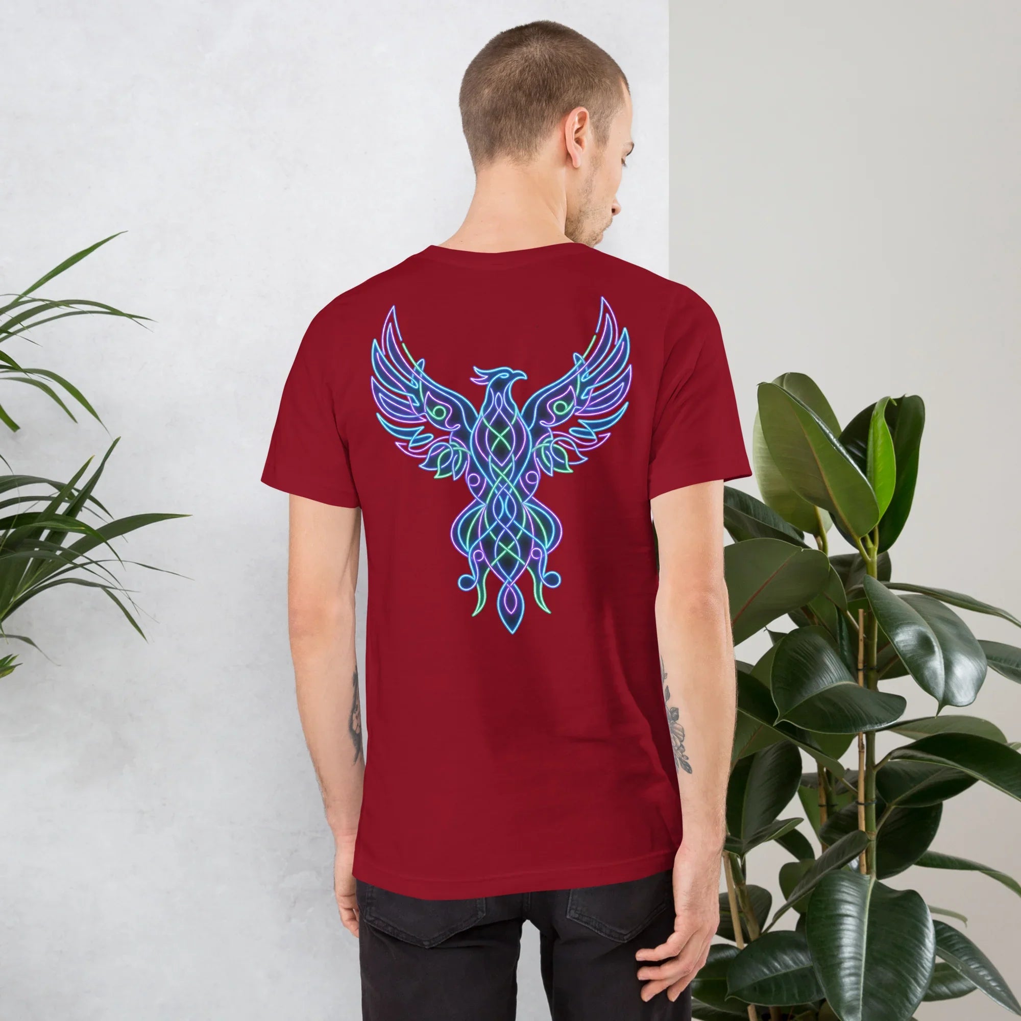 Man wearing cardinal red t-shirt with neon blue and green phoenix design on back in indoor setting with plants
