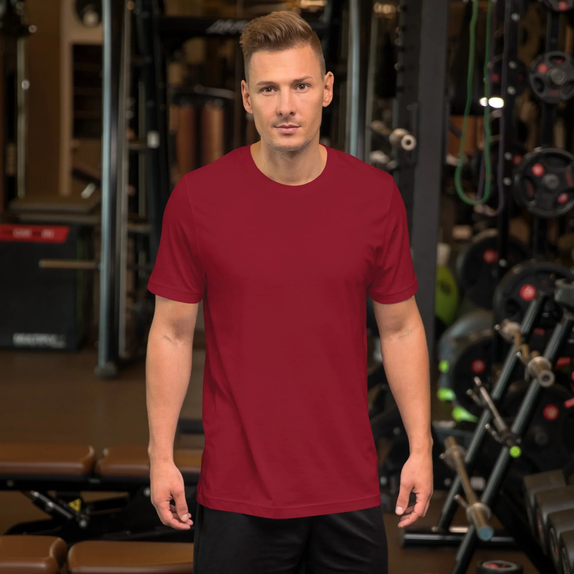 Man wearing a plain cardinal red unisex staple t-shirt in a gym setting with workout equipment