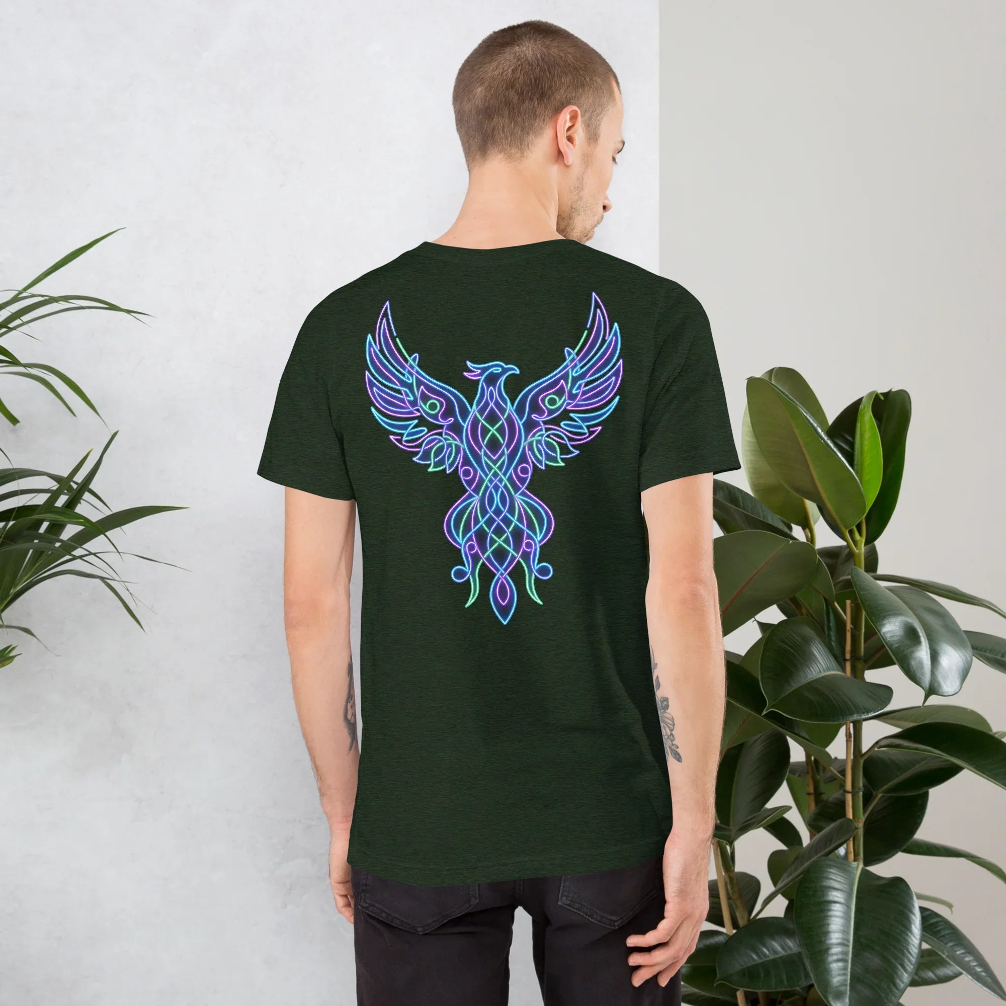 Back view of a man wearing a dark green t-shirt with a vibrant neon phoenix graphic, standing indoors by green plants