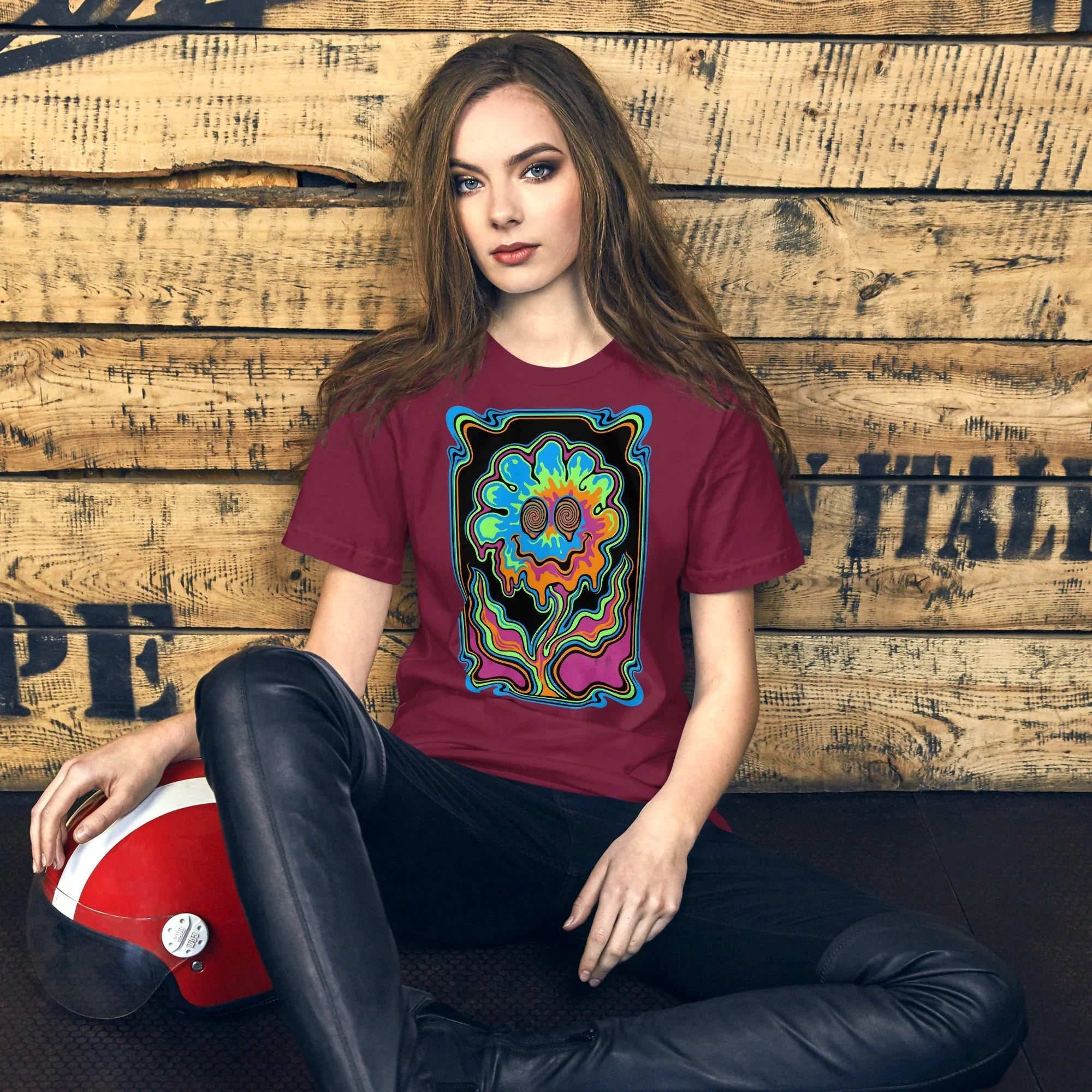 Woman wearing maroon psychedelic graphic t-shirt with colorful melting flower design, sitting against wood wall holding red helmet
