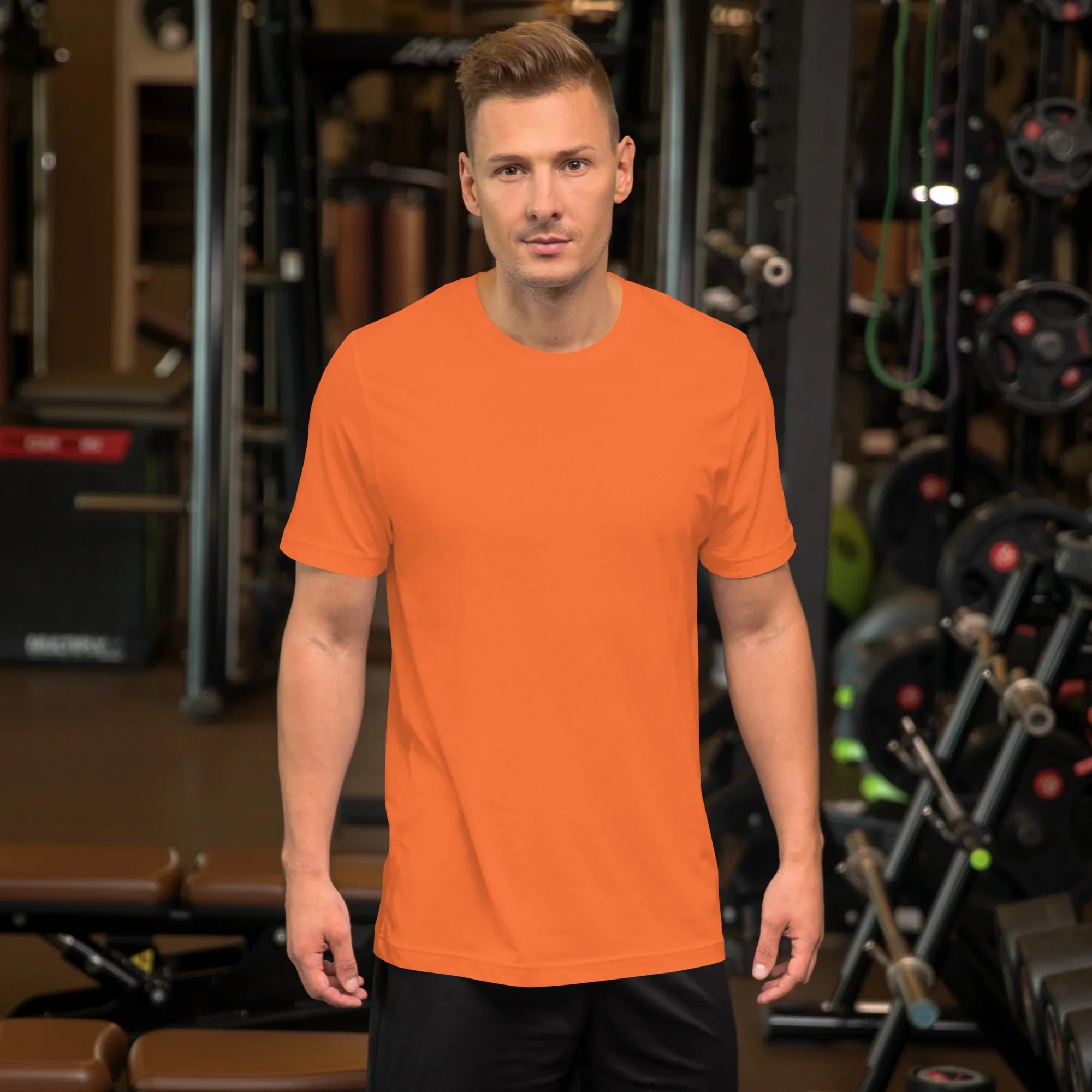 Man wearing plain orange unisex staple t-shirt standing in gym with workout equipment