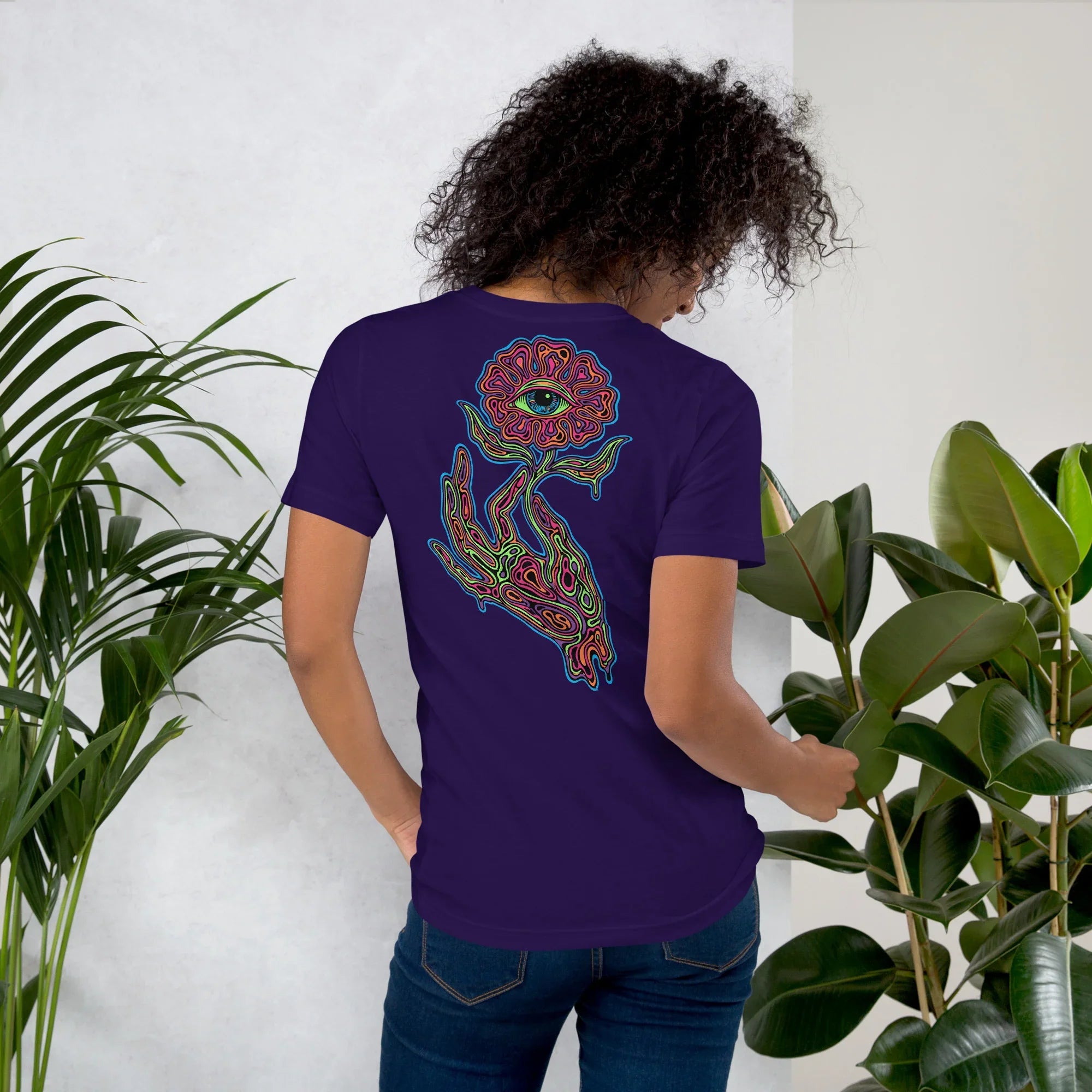 Woman wearing purple unisex t-shirt with psychedelic hand holding eye-flower design among green plants