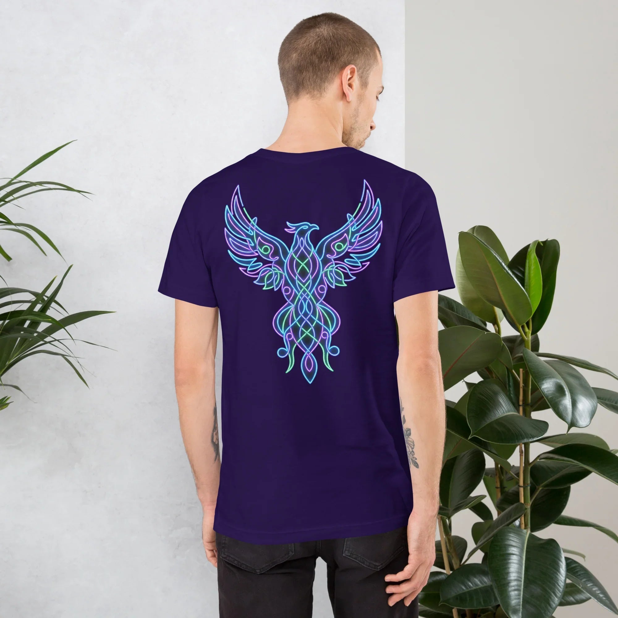 Man wearing unisex staple purple t-shirt with neon phoenix design on back, standing near green plants