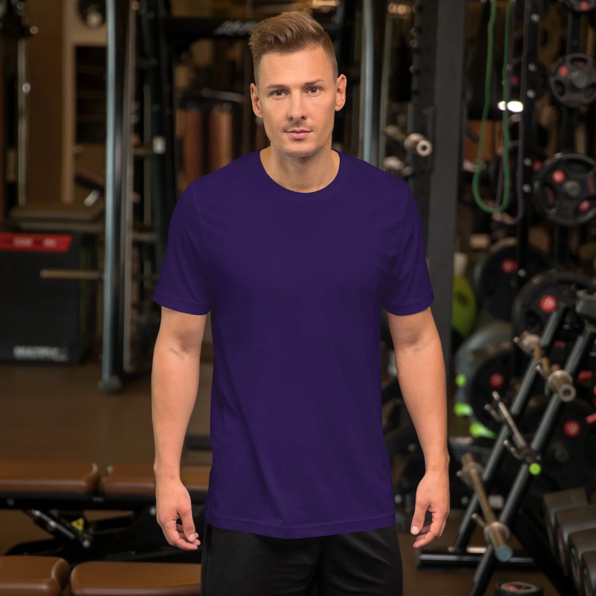 Man wearing a plain unisex purple staple t-shirt standing in a gym with workout equipment in the background