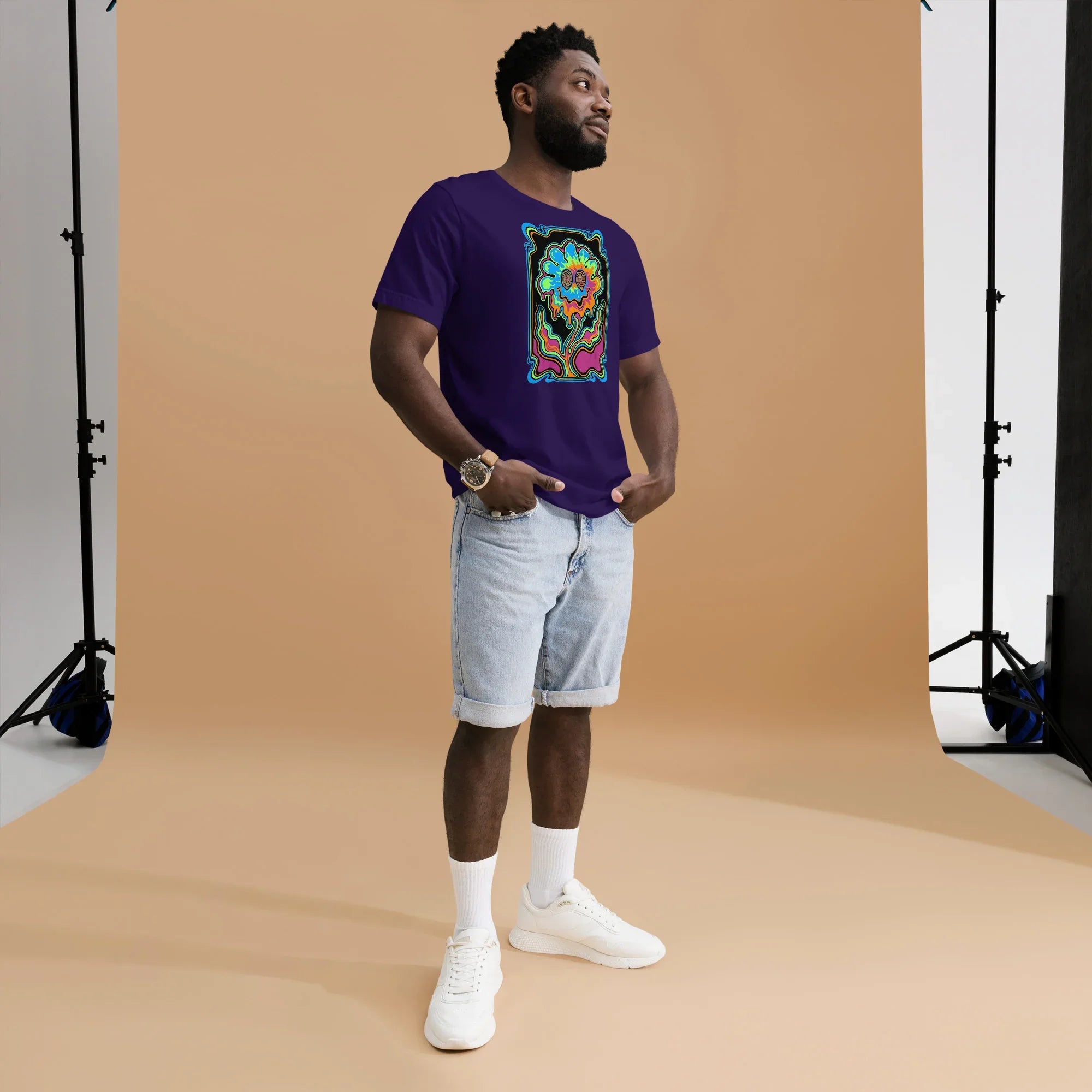 Man wearing a unisex purple t-shirt with vibrant psychedelic flower graphic, light denim shorts, white sneakers, studio backdrop
